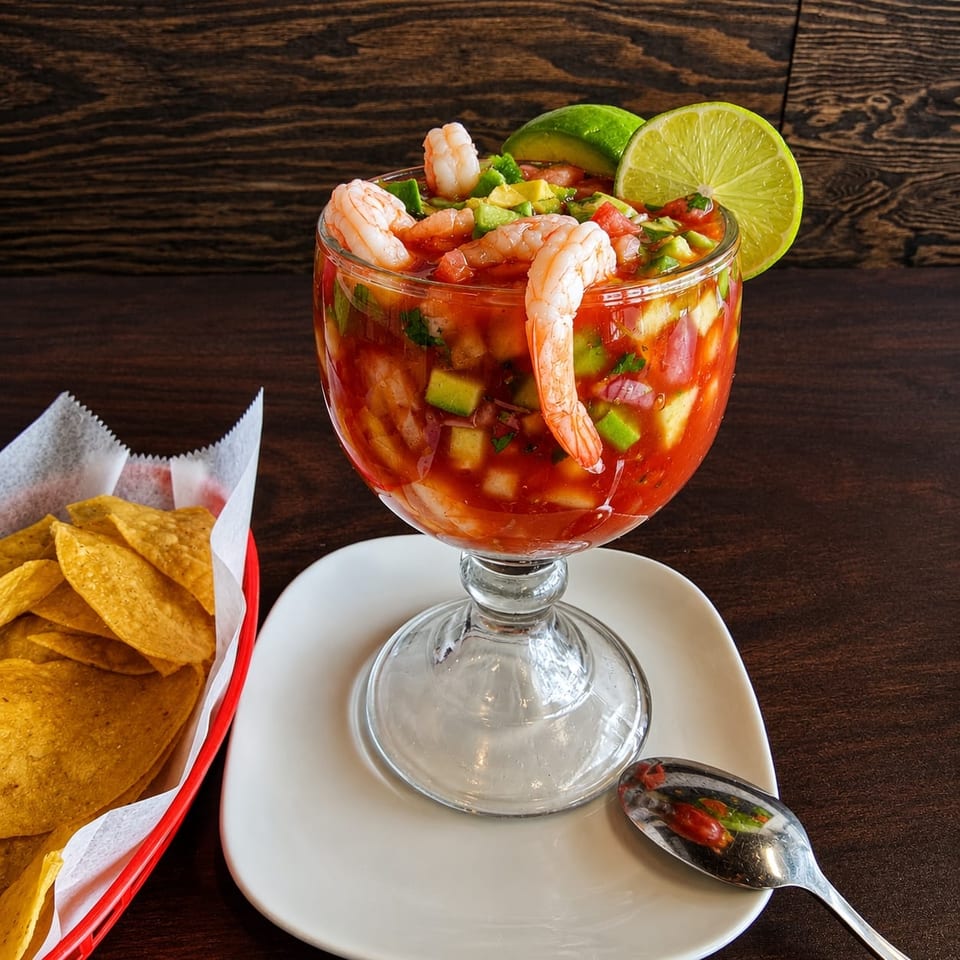 Shrimp ceviche with diced vegetables and lime garnish in a glass goblet, served with tortilla chips on a white plate.
