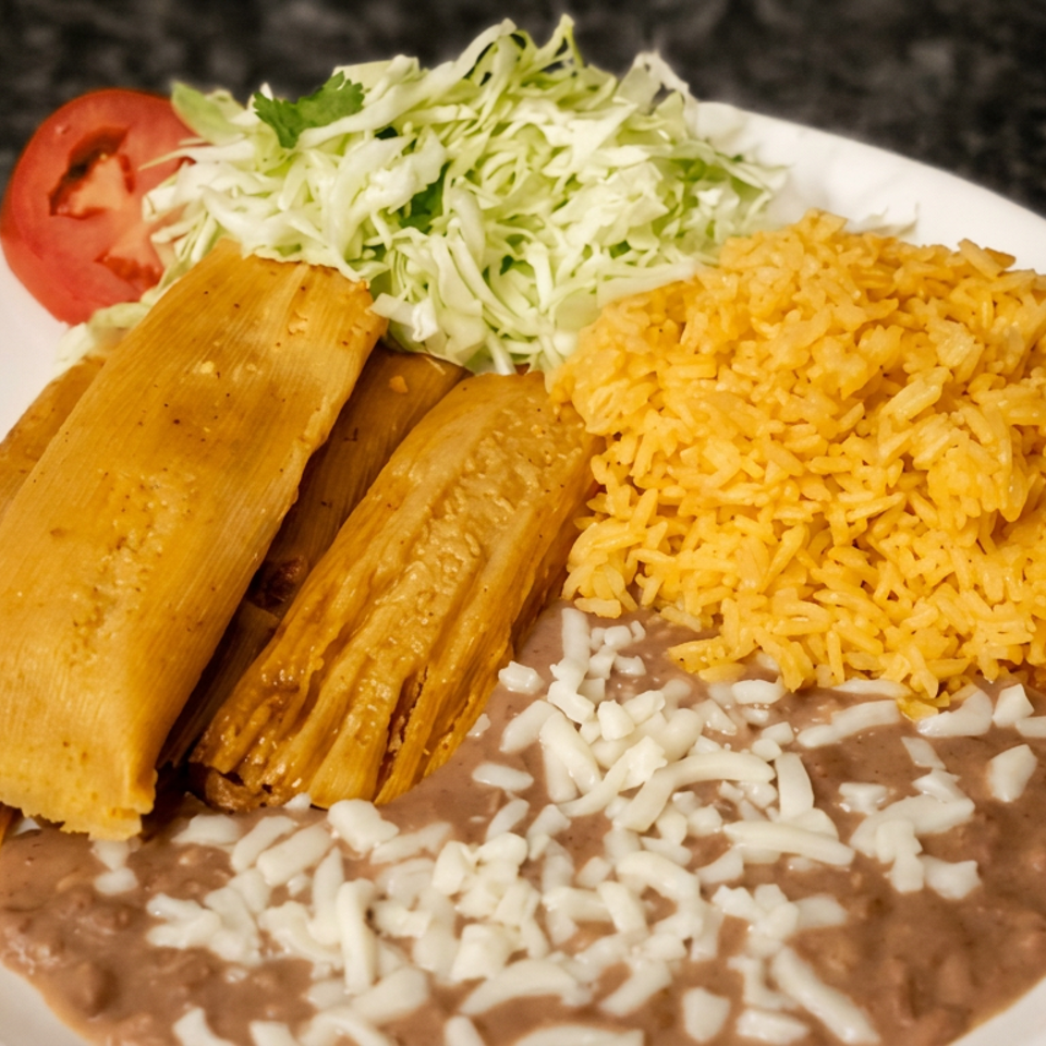 Mexican plate with two tamales, yellow rice, refried beans topped with cheese, and cabbage salad with tomato.